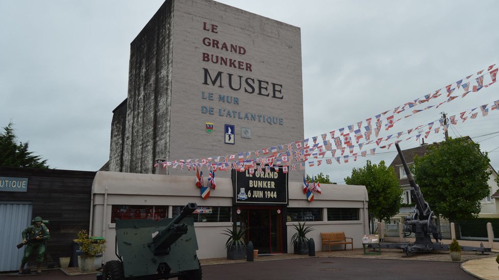 Exterior of the Grand Bunker Museum in Ouistreham, showing the tall concrete bunker with artillery and a Flak 88 gun on display